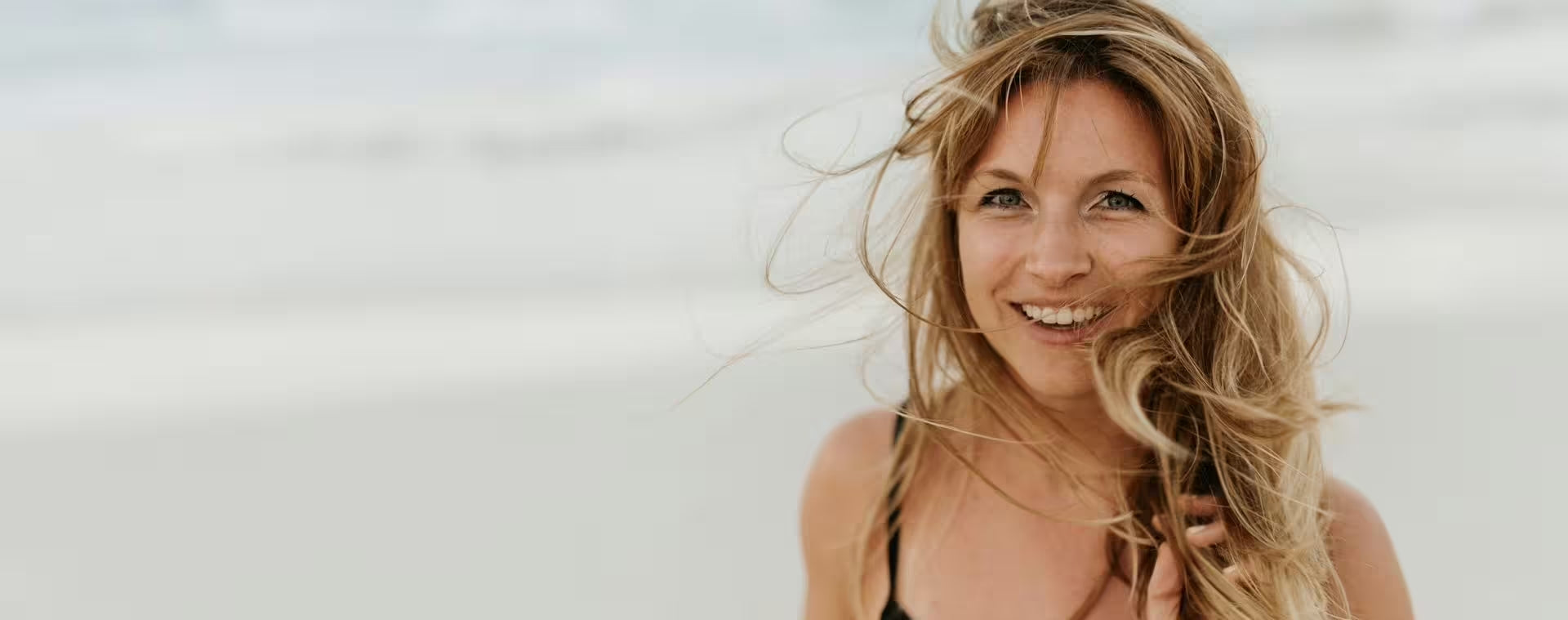 Eine Frau mit langen, hellbraunen Haaren lächelt an einem Sandstrand in die Kamera, wobei ihr Haar im Wind weht. Der Hintergrund ist sanft unscharf und erzeugt so eine leichte und luftige Atmosphäre.