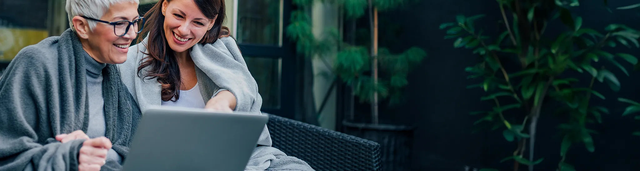 Zwei Frauen sitzen lächelnd mit einem Laptop zusammen, in Decken gehüllt. Der Text auf Deutsch lautet: "Angebote entdecken und Rabatte sichern".