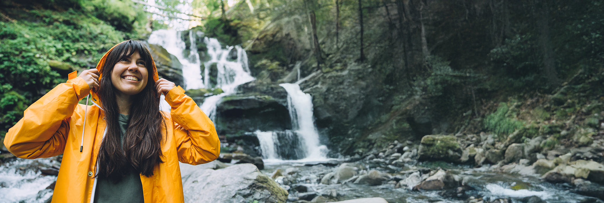 Eine lächelnde Frau in einer orangefarbenen Regenjacke steht in der Nähe eines felsigen Baches und Wasserfalls in einem üppigen, grünen Wald.