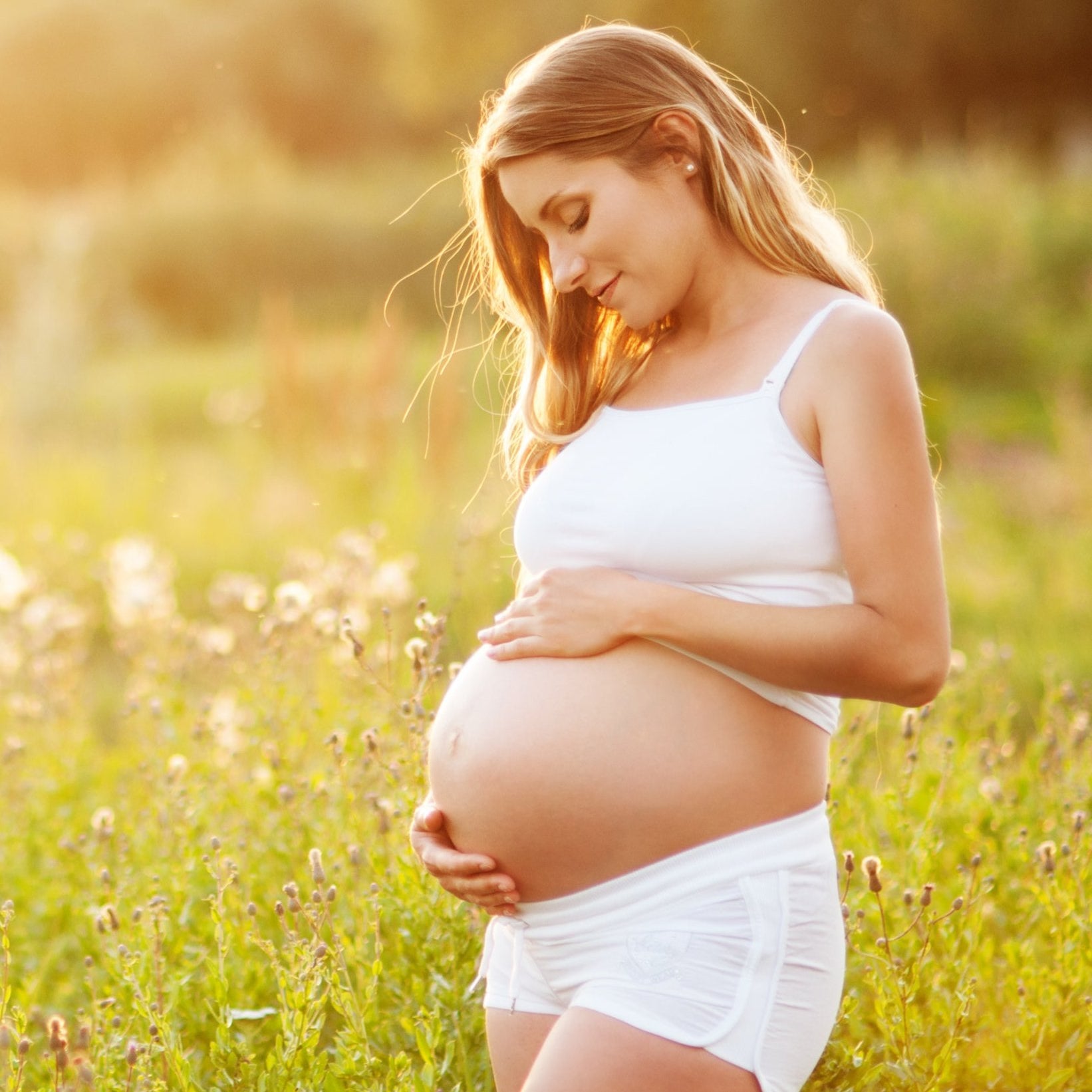 Eine schwangere Frau in weißem Tanktop und Shorts steht auf einer sonnenbeschienenen Wiese, wiegt sanft ihren Bauch und blickt mit einem sanften Lächeln nach unten. Wildblumen und Grün umgeben sie im warmen, goldenen Licht.