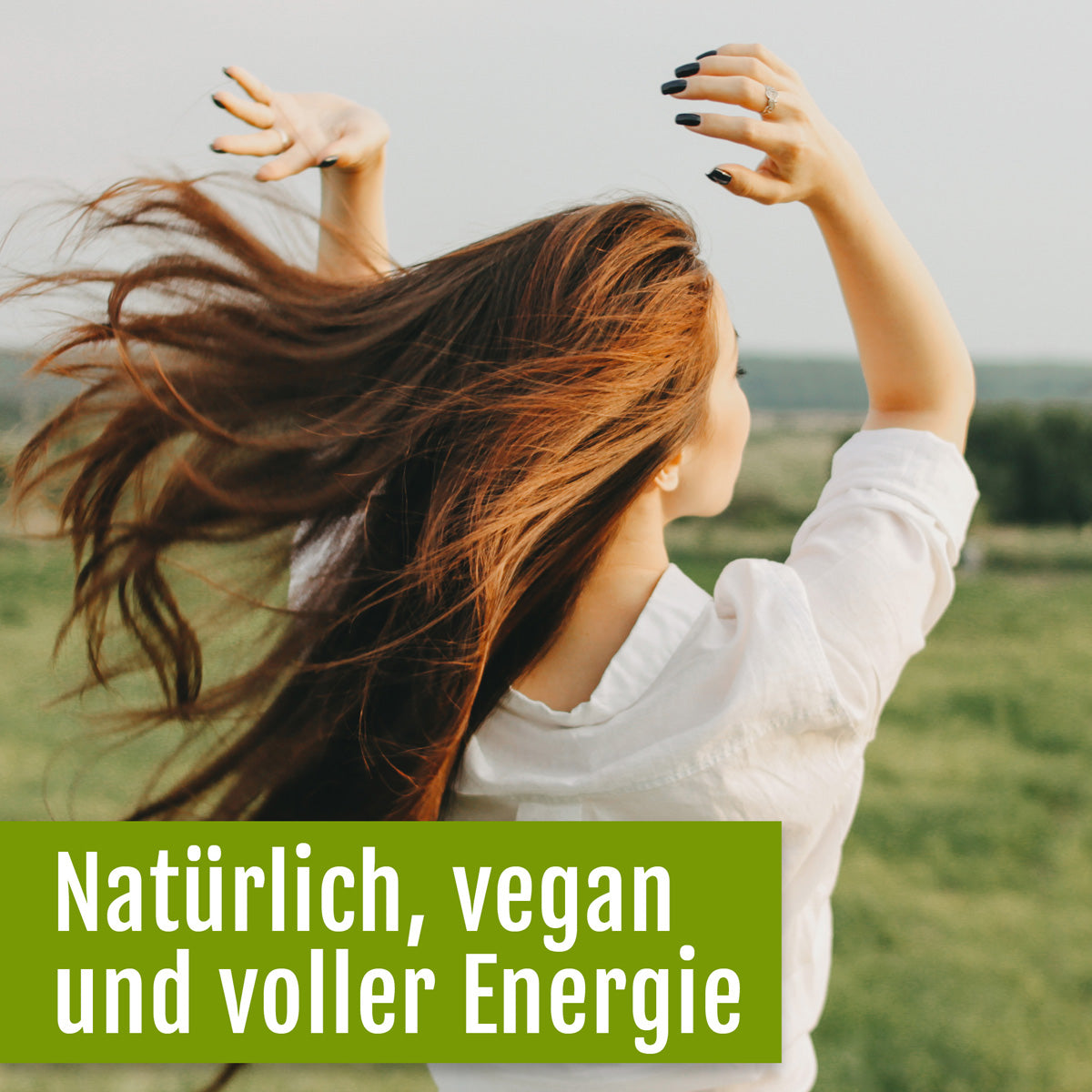Eine fröhliche Frau mit langen braunen Haaren und einem weißen Hemd streckt auf einer grünen Wiese die Arme in die Höhe. Deutscher Bildtext: Natürlich, vegan und voller Energie – dank Naturland-Bio-Chlorella-Tabletten von gse.organic.supplements und wertvollem Vitamin B12.