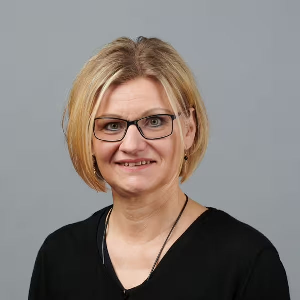 Eine Frau mit kurzen blonden Haaren und Brille, die ein schwarzes Oberteil und eine Halskette trägt, lächelt vor einem schlichten grauen Hintergrund.