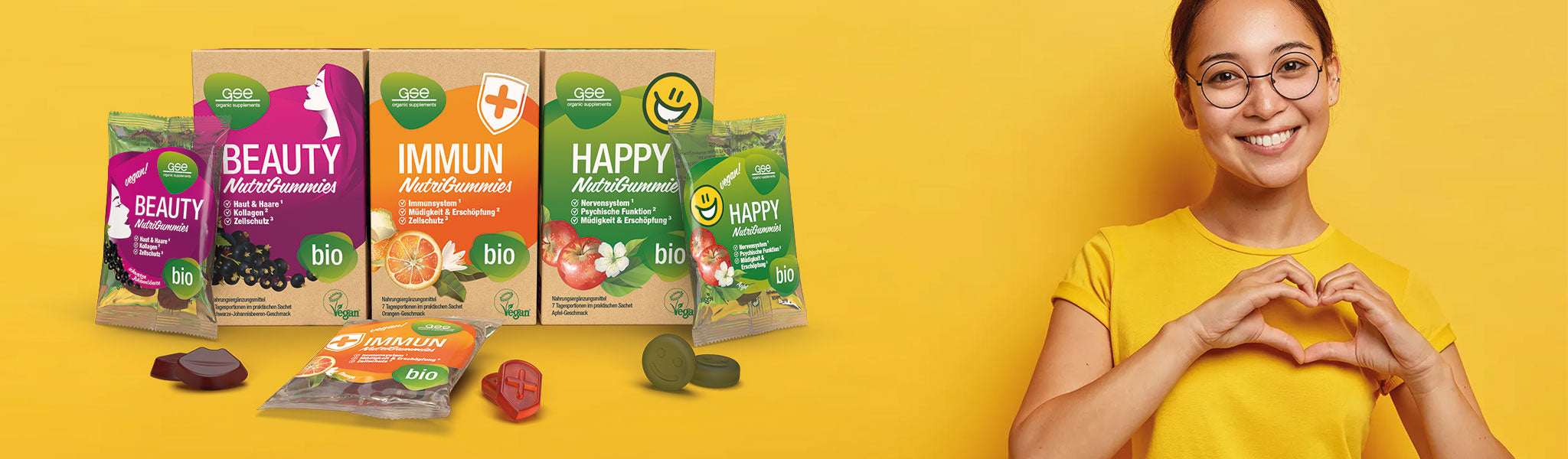 Eine lächelnde Frau mit Brille und gelbem Shirt formt mit ihren Händen ein Herz. Neben ihr liegen Packungen mit „Bio Nutri Gummies“ mit den Aufschriften „Beauty“, „Immun“ und „Happy“, auf denen mehrere Gummibärchen zu sehen sind.