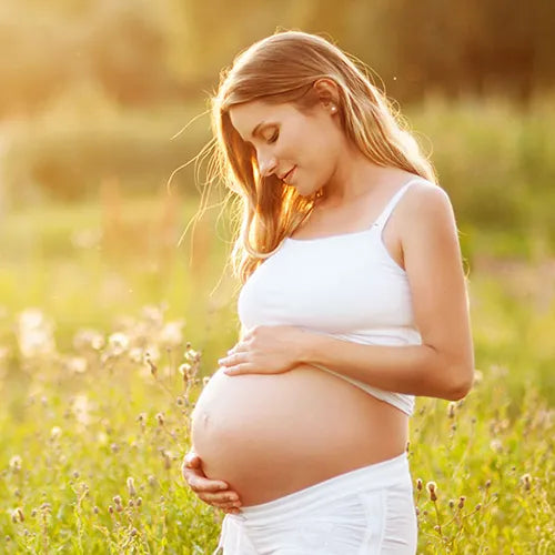 Eine schwangere Frau in einem weißen Outfit steht auf einem sonnenbeschienenen Feld, wiegt sanft ihren Bauch und lächelt, während sie nach unten blickt, umgeben von grünem Gras und Wildblumen.