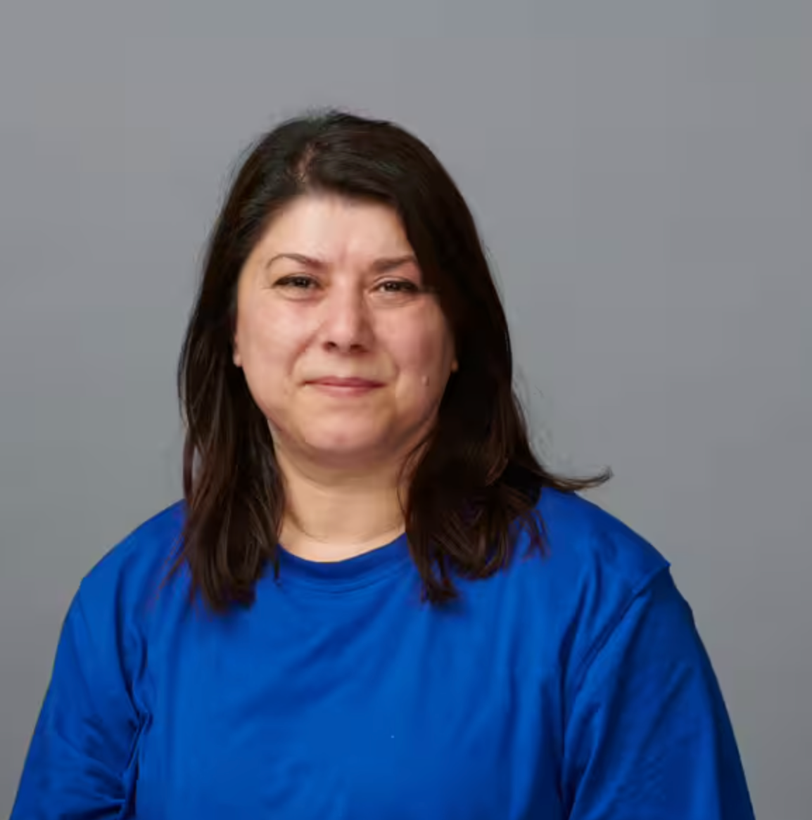 Eine Frau mit mittellangem dunklem Haar und einem leuchtend blauen T-Shirt sitzt vor einem schlichten grauen Hintergrund und blickt mit einem subtilen Lächeln in die Kamera.