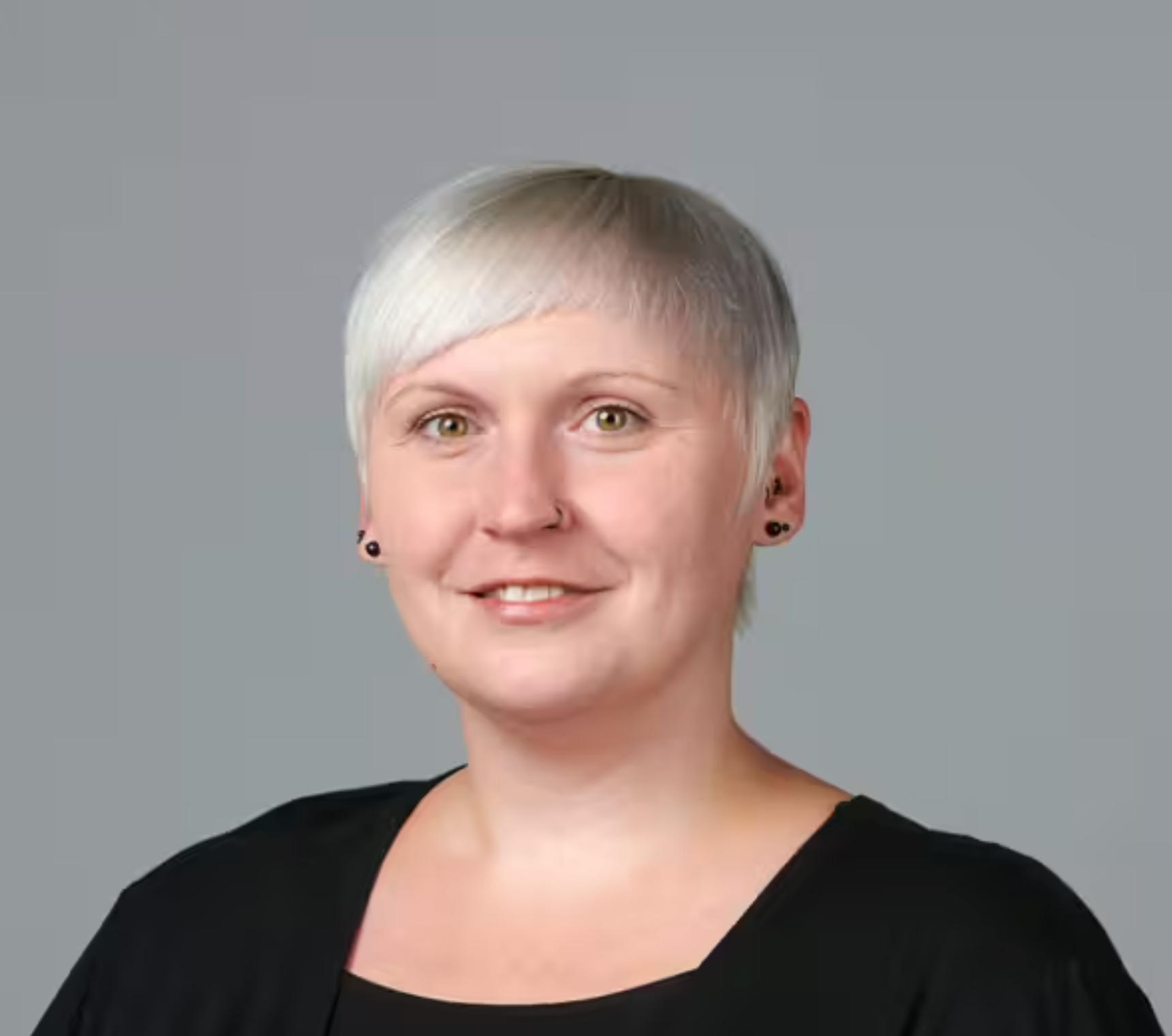 Eine lächelnde Person mit kurzem, glattem, platinblondem Haar, die ein schwarzes Oberteil und kleine schwarze Ohrringe trägt und vor einem schlichten grauen Hintergrund posiert.