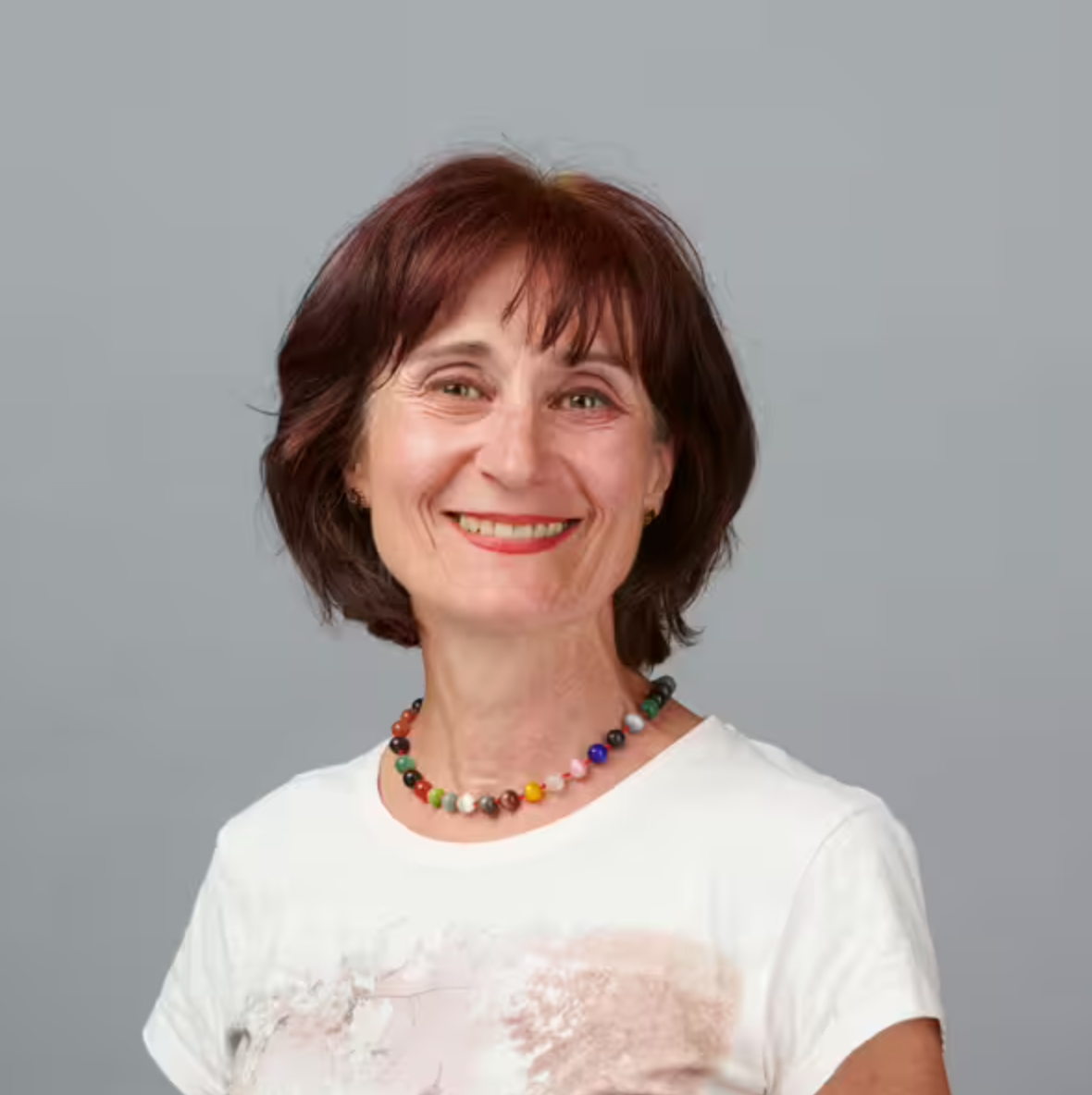 Eine Frau mit kurzen braunen Haaren lächelt in die Kamera. Sie trägt ein weißes T-Shirt und eine bunte Perlenkette. Der Hintergrund ist schlicht hellgrau.
