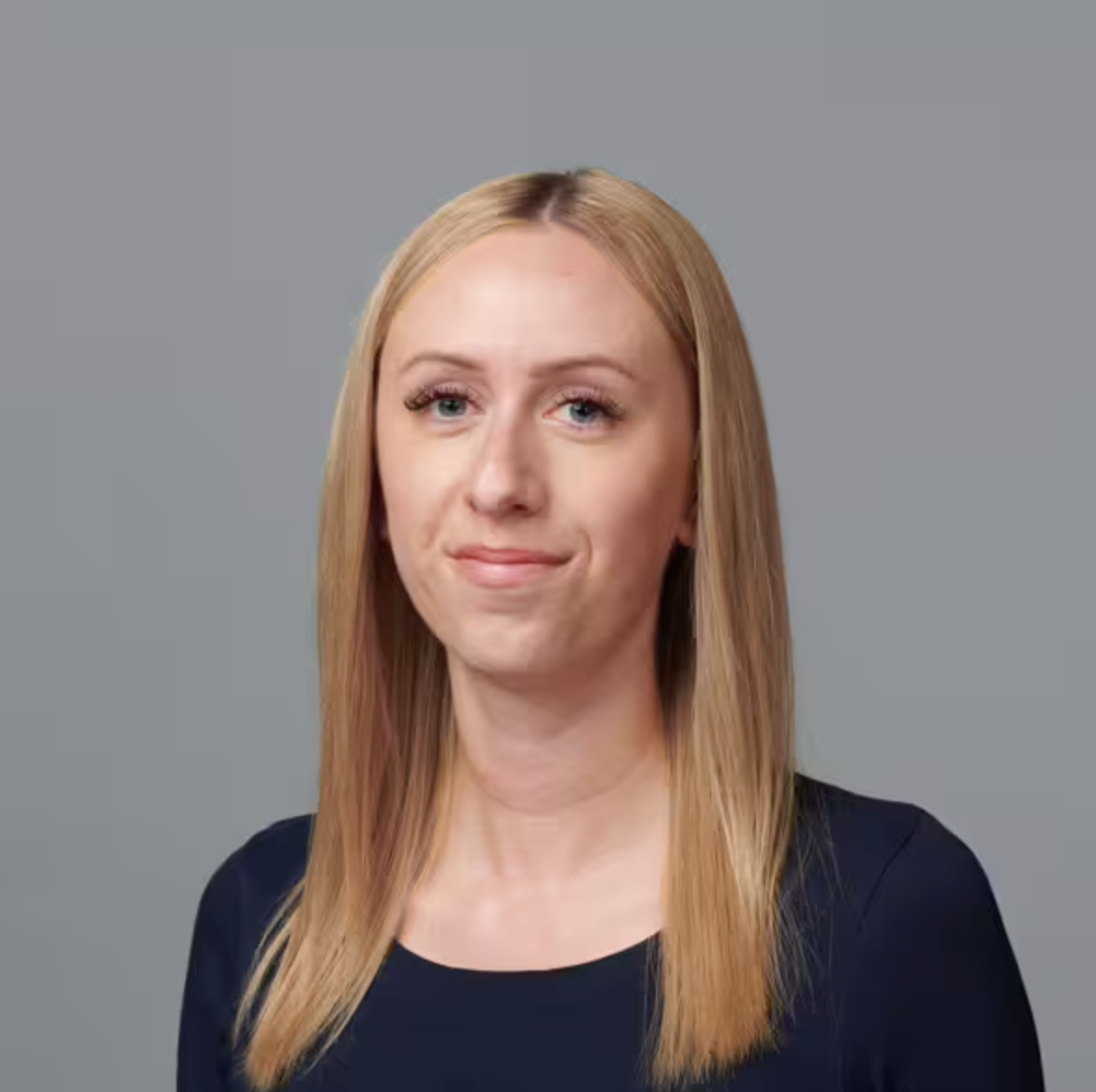 Eine Frau mit glattem, schulterlangem blondem Haar und heller Haut trägt ein dunkelblaues Oberteil. Sie blickt nach vorne und lächelt leicht vor einem schlichten grauen Hintergrund.