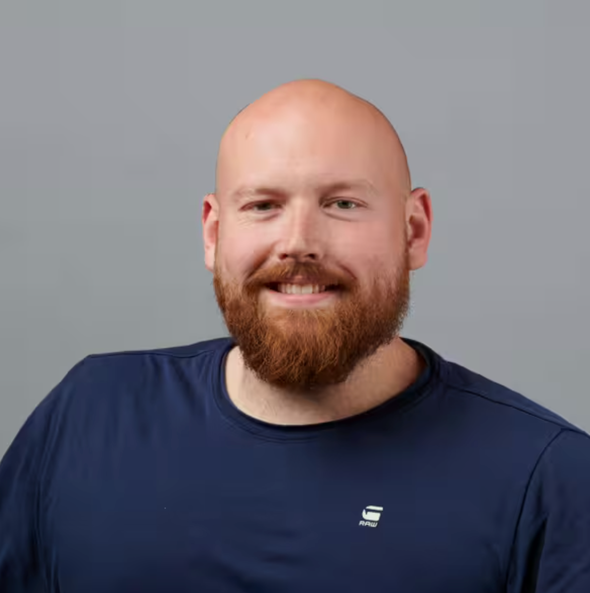 Ein bärtiger Mann mit rasiertem Kopf lächelt in die Kamera. Er trägt ein marineblaues T-Shirt und posiert vor einem schlichten grauen Hintergrund.
