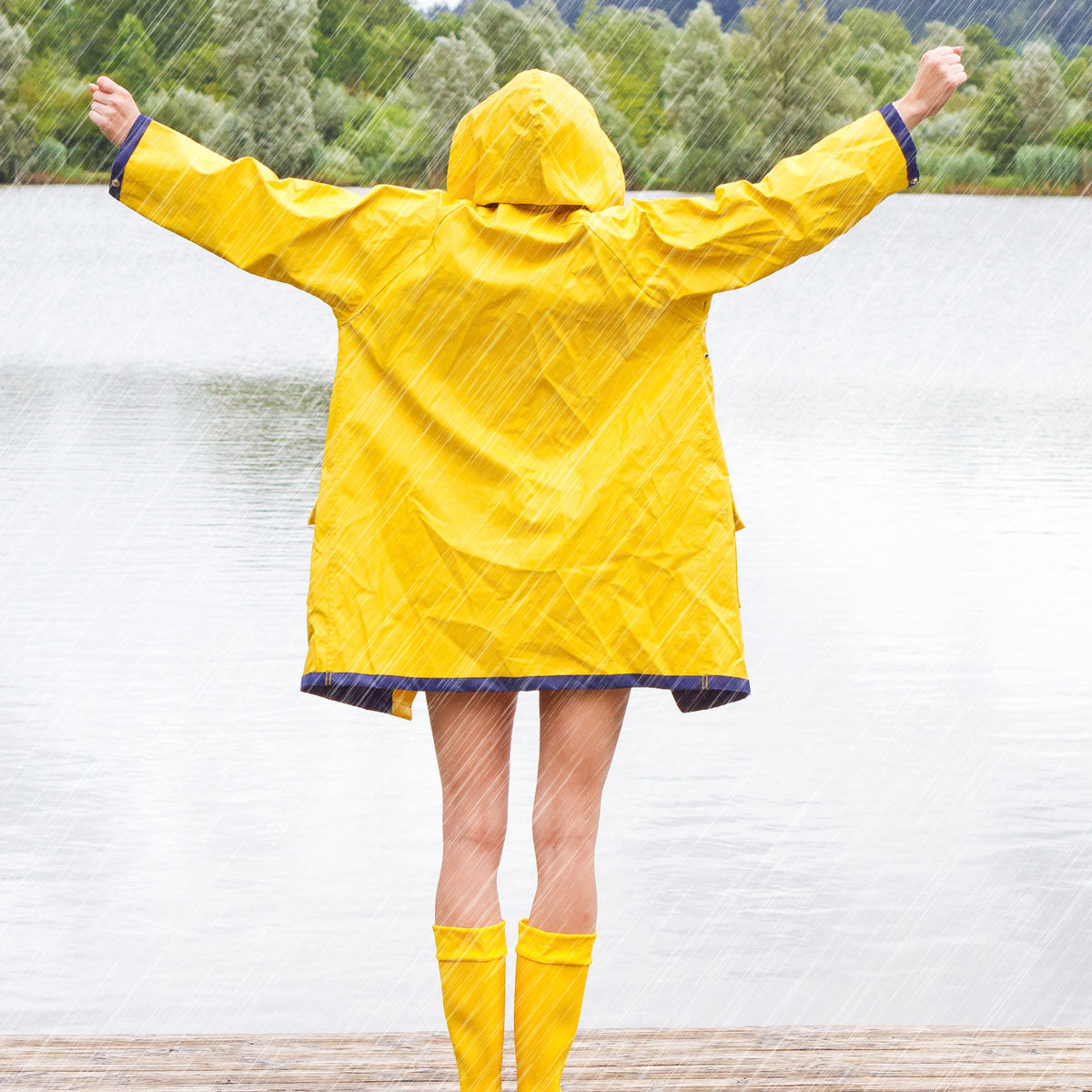 Eine Person in einem gelben Regenmantel und Stiefeln steht mit ausgestreckten Armen im Regen auf einem Holzsteg und blickt auf einen See mit Bäumen und Hügeln im Hintergrund.