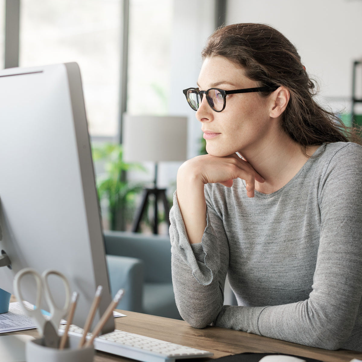 Eine Frau mit Brille und zurückgebundenem langen braunen Haar sowie einem grauen Langarmhemd sitzt an einem Schreibtisch und schaut aufmerksam auf einen Computermonitor in einem hellen, modernen Büro.