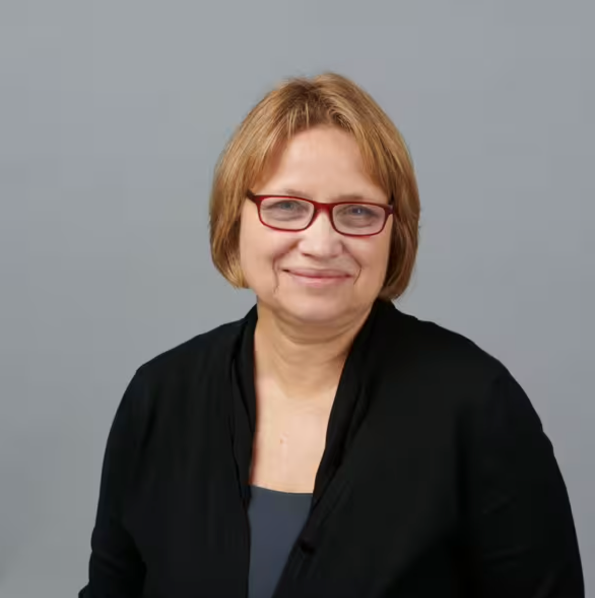 Eine Frau mittleren Alters mit kurzen hellbraunen Haaren, einer roten Brille, einer schwarzen Strickjacke und einem grauen Oberteil lächelt sanft vor einem schlichten grauen Hintergrund.