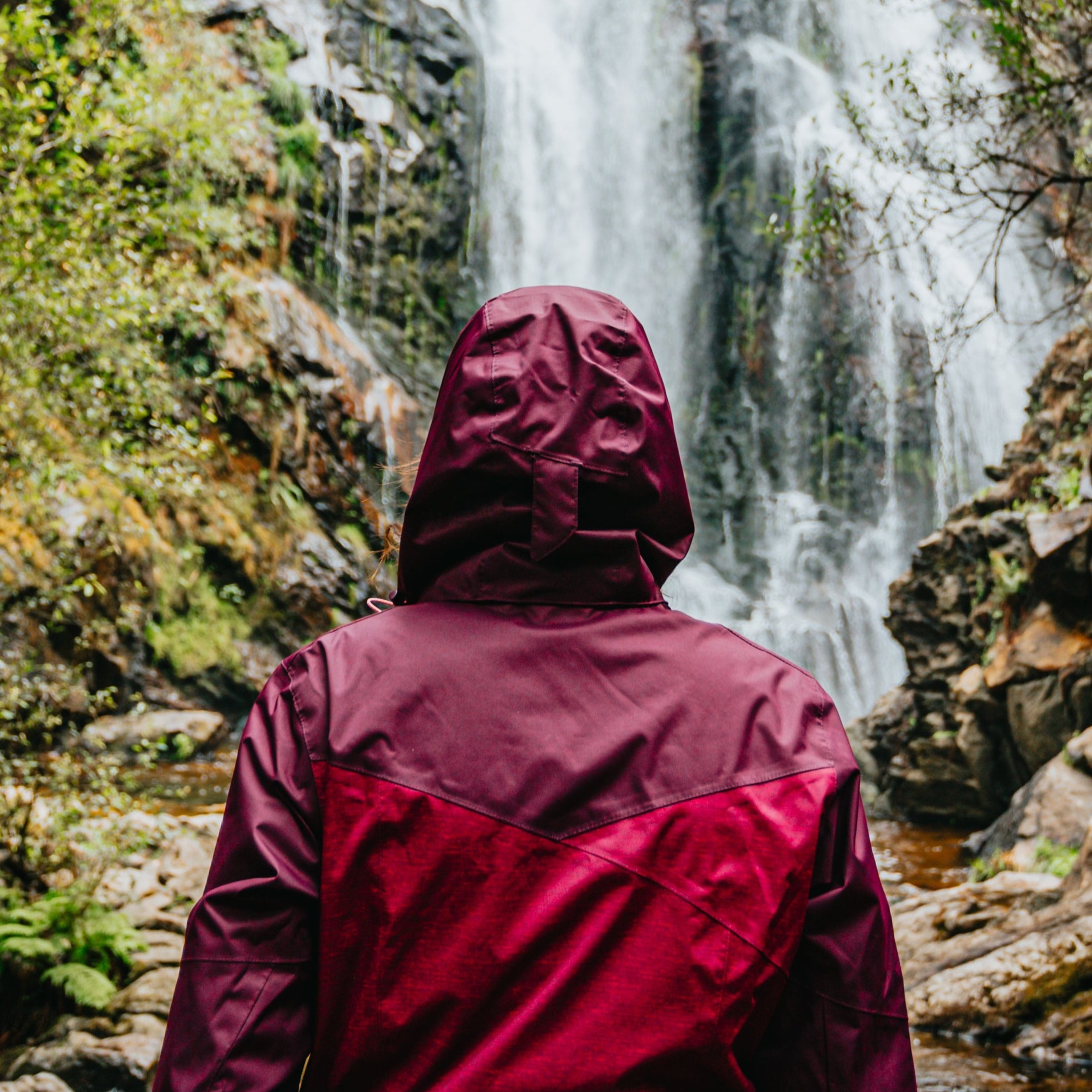 Eine Person in einer kastanienbraunen Kapuzenjacke steht mit dem Rücken zur Kamera und blickt auf einen hohen Wasserfall, der von Felsklippen und viel Grün umgeben ist.