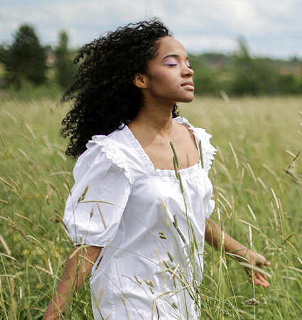 Eine Frau mit lockigem Haar und einem weißen Kleid geht mit geschlossenen Augen durch eine hohe Grasfläche und sieht unter einem teilweise bewölkten Himmel friedlich und entspannt aus.