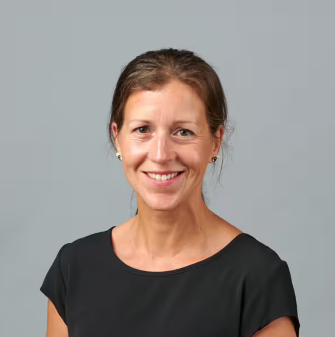 Eine Frau mit zurückgekämmtem braunem Haar, einem schwarzen Kurzarmoberteil und kleinen Ohrringen lächelt vor einem schlichten grauen Hintergrund in die Kamera.