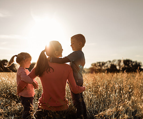 Eine Frau mit Pferdeschwanz sitzt auf einer sonnenbeschienenen Wiese und hält einen kleinen Jungen im Arm. Ein kleines Mädchen steht daneben. Die Sonne steht tief am Himmel und hüllt die Familie in einen warmen, goldenen Schein.