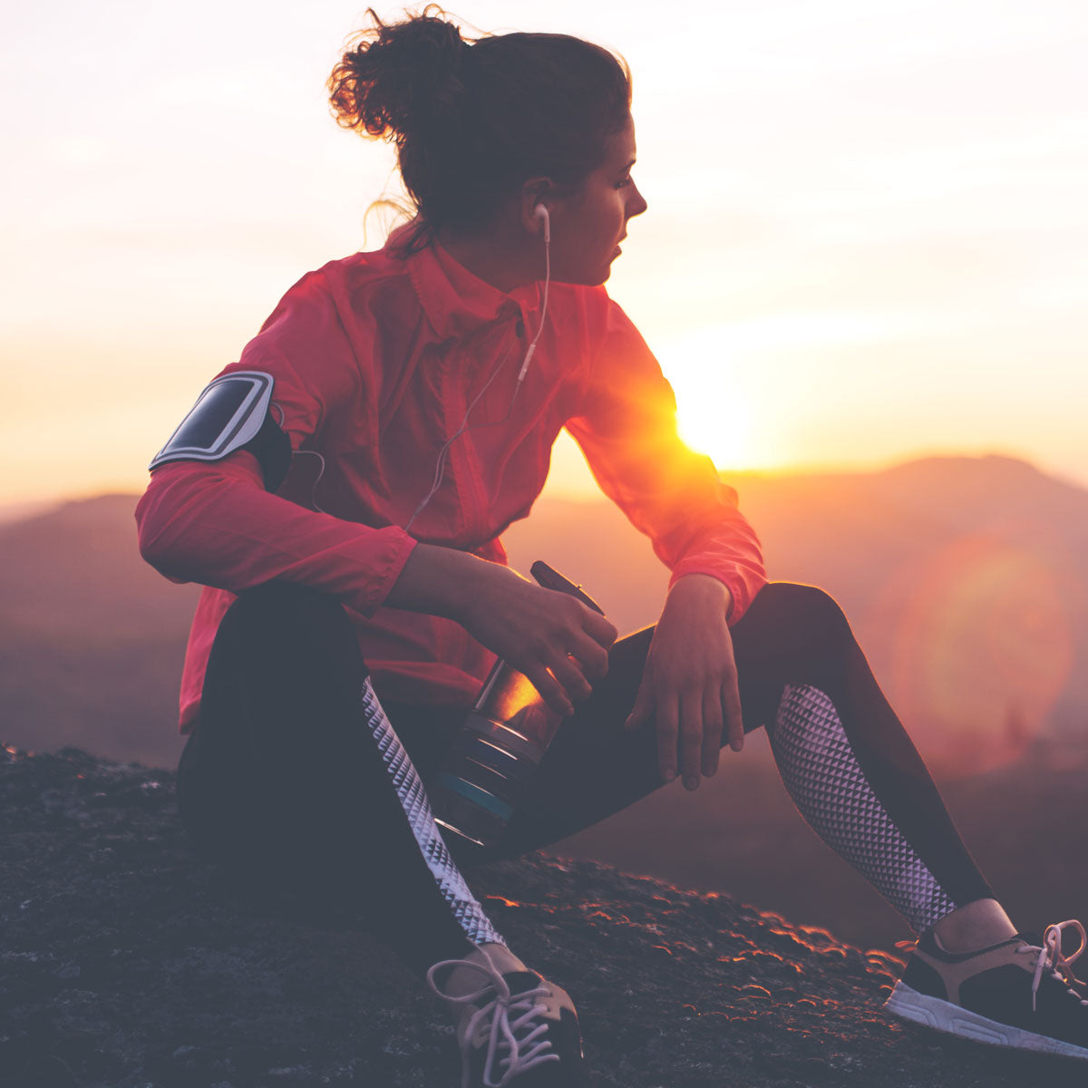 Eine Frau in Sportkleidung sitzt bei Sonnenuntergang auf einem Felsen, hält eine Wasserflasche in der Hand und trägt Kopfhörer und eine Armbinde, während sie auf die Berge in der Ferne blickt.