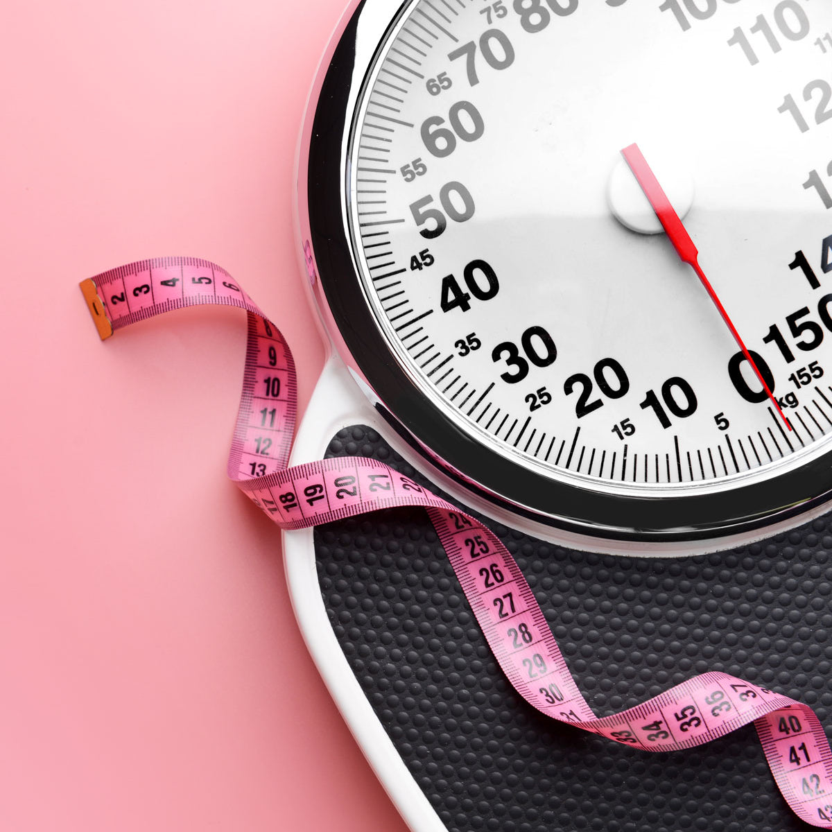 Eine Personenwaage mit schwarzer Plattform und Zifferblattanzeige steht auf einem rosa Hintergrund neben einem aufgerollten rosa Maßband.