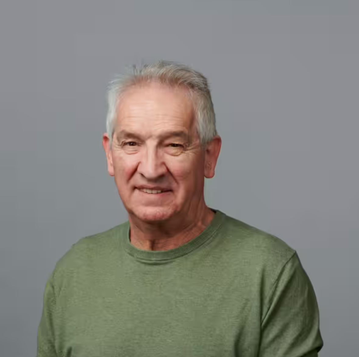 Ein älterer Mann mit kurzen grauen Haaren und einem grünen Pullover sitzt vor einem schlichten grauen Hintergrund und lächelt sanft in die Kamera.